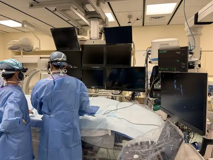 Two operators wearing VistaMD augmented reality headsets work from virtual screens at the procedure table. They both see the same images, although they can distribute them differently. The boom monitor is off.